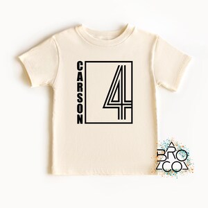 May include: A cream-colored short-sleeve t-shirt featuring a black graphic design on the front. The design consists of a rectangular border containing the name CARSON written vertically in black capital letters next to a large black number 4.