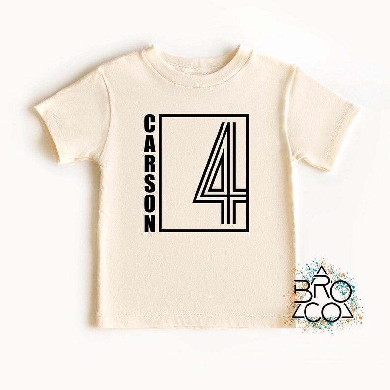 May include: A cream-colored short-sleeve t-shirt featuring a black graphic design on the front. The design consists of a rectangular border containing the name CARSON written vertically in black capital letters next to a large black number 4.