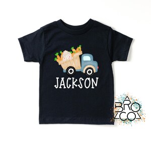 May include: A black short-sleeved t-shirt with a cartoon illustration of a blue truck carrying a basket of carrots and a bunny. The text "JACKSON" is printed in white below the truck.