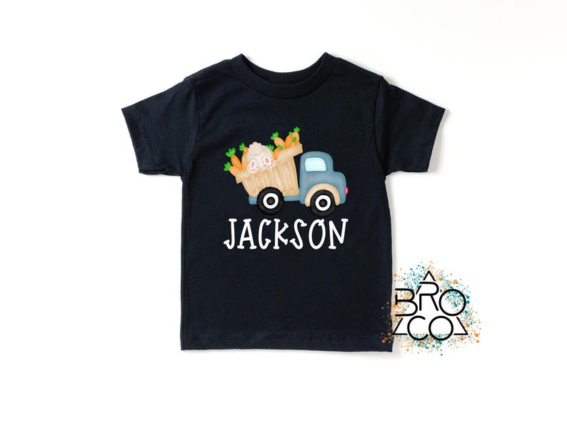 May include: A black short-sleeved t-shirt with a cartoon illustration of a blue truck carrying a basket of carrots and a bunny. The text "JACKSON" is printed in white below the truck.