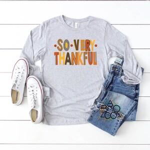 May include: A light gray long-sleeve t-shirt featuring the text SO VERY THANKFUL in multicolored block letters, paired with blue denim jeans and white canvas sneakers. The shirt has a crew neckline and the jeans have a classic five-pocket design.