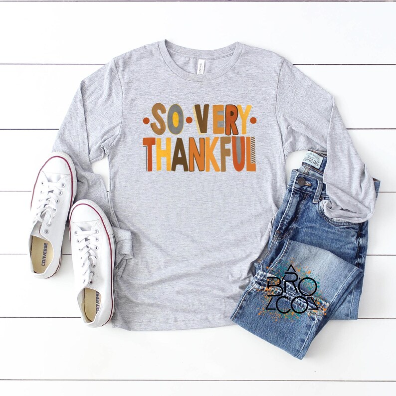 May include: A light gray long-sleeve t-shirt featuring the text SO VERY THANKFUL in multicolored block letters, paired with blue denim jeans and white canvas sneakers. The shirt has a crew neckline and the jeans have a classic five-pocket design.
