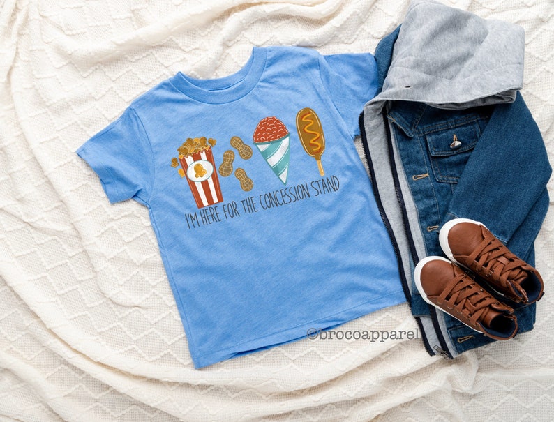 May include: A light blue t-shirt featuring a graphic of popcorn, peanuts, a snow cone, and a corn dog. The text "I'm here for the concession stand" is printed on the shirt.