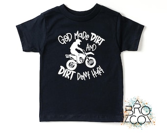 Könnte beinhalten: Ein schwarzes kurzärmeliges T-Shirt mit einer weißen Grafik einer Person auf einem Dirtbike. Der in Weiß gedruckte Text lautet: God Made Dirt And Dirt Don't Hurt. Das Design ist mittig auf der Brust des T-Shirts platziert und hat einen Rundhalsausschnitt.