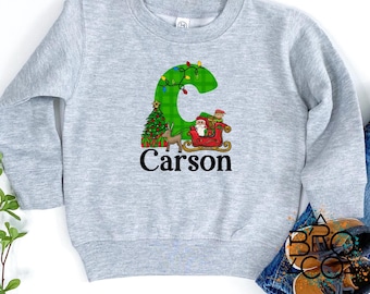 May include: A light gray sweatshirt featuring a large green letter "C" decorated with Christmas lights. The letter has a Christmas tree and Santa Claus in a sleigh. The name "Carson" is printed below the letter.