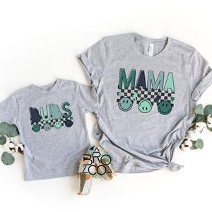 May include: Grey t-shirts with a chequered pattern and smiley faces. The larger shirt says "MAMA" and the smaller shirt says "DIBS".