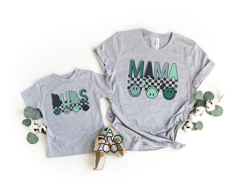 May include: Grey t-shirts with a chequered pattern and smiley faces. The larger shirt says "MAMA" and the smaller shirt says "DIBS".