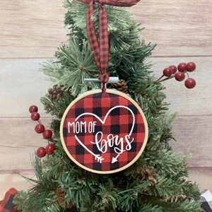 May include: A Christmas tree ornament with a red and black buffalo plaid design. The ornament has the words "MOM OF BOYS" in white script inside a heart shape. A red and black plaid ribbon is attached for hanging.