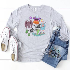 May include: A pale grey long-sleeved top with a vibrant graphic that reads "I BELIEVE." The design includes a purple UFO, a winged creature, a blue monster, a green sea serpent, and other fantastical elements. White trainers and blue jeans are also visible.