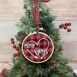 May include: A Christmas tree ornament with a red and black buffalo plaid design. The ornament has the words "MOM OF BOYS" in white script inside a heart shape. A red and black plaid ribbon is attached.