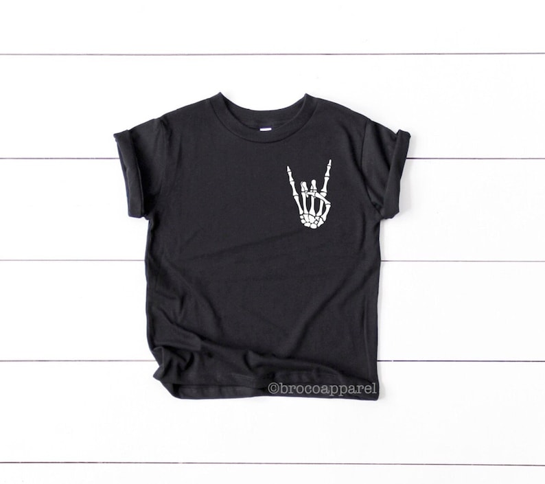 May include: Black t-shirt with a white skeleton hand making a rock and roll sign on the front. The shirt has rolled up sleeves.