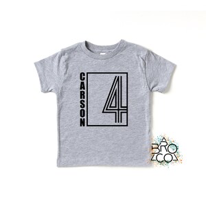 May include: A heather gray short-sleeve t-shirt featuring a black graphic design on the front. The design includes the name CARSON written vertically next to a large outlined number 4 inside a rectangular frame. A small logo reads BRO CO.