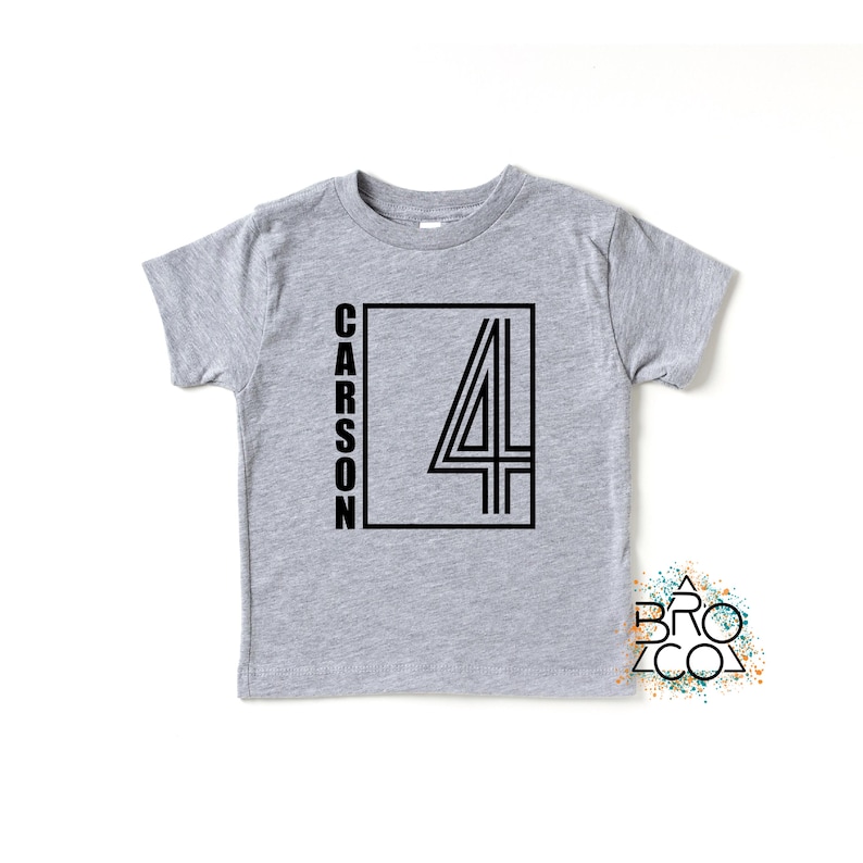 May include: A heather gray short-sleeve t-shirt featuring a black graphic design on the front. The design includes the name CARSON written vertically next to a large outlined number 4 inside a rectangular frame. A small logo reads BRO CO.