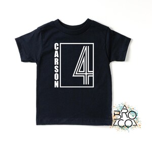 May include: A black short-sleeved t-shirt featuring a white graphic design on the front. The design includes a rectangular frame containing the number 4 and the name CARSON written vertically in white capital letters along the left side.