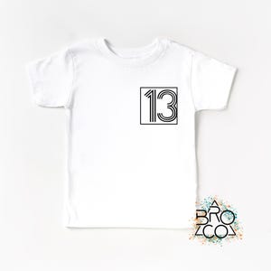 Op de afbeelding: Wit T-shirt met het zwarte nummer 13 in een vierkant. Het nummer is ontworpen met verticale lijnen. Een eenvoudige, casual kledingstijl.