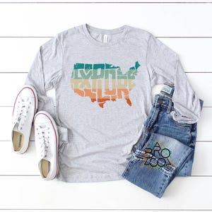 May include: A heather gray long-sleeve t-shirt featuring a graphic of the United States map with the word EXPLORE printed inside in a retro-style font with teal, yellow, and orange horizontal stripes, paired with white sneakers and blue jeans.