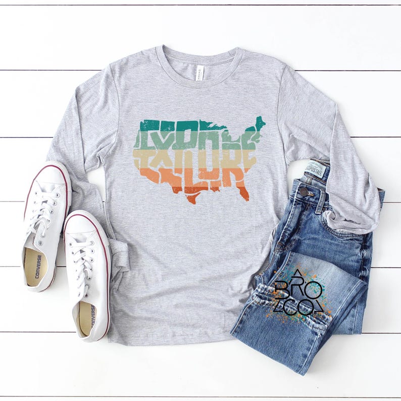 May include: A heather gray long-sleeve t-shirt featuring a graphic of the United States map with the word EXPLORE printed inside in a retro-style font with teal, yellow, and orange horizontal stripes, paired with white sneakers and blue jeans.