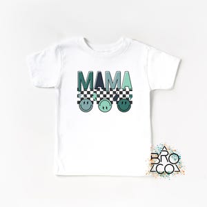 May include: White t-shirt with the word "MAMA" in green and blue letters with a checkered pattern and three green and blue smiley faces underneath.