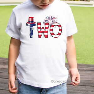Op de afbeelding: Een wit T-shirt met het woord "TWEE" in rood, wit en blauw, met een rood, wit en blauwe hoed en vuurwerk bovenop de letters.