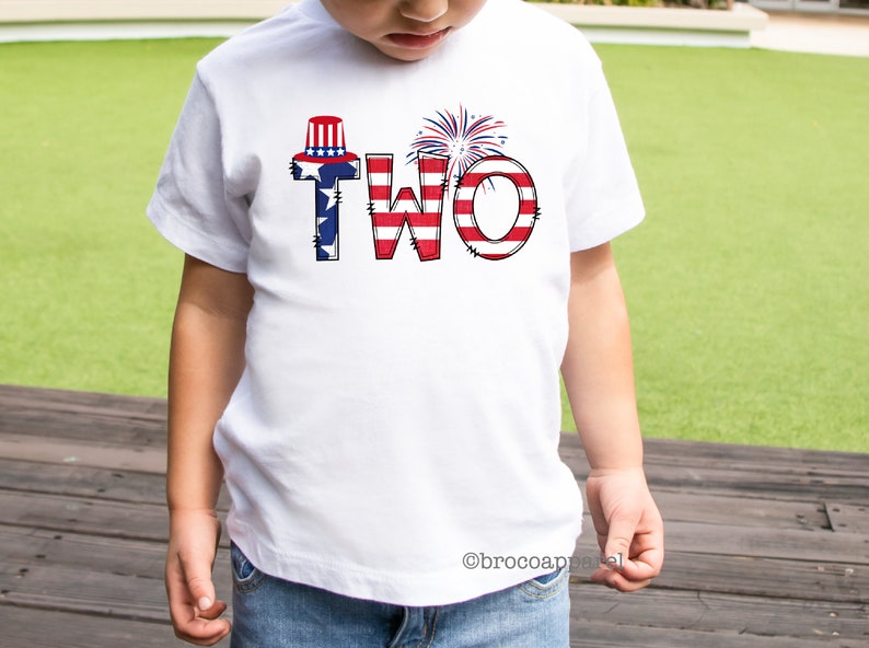 Op de afbeelding: Een wit T-shirt met het woord "TWEE" in rood, wit en blauw, met een rood, wit en blauwe hoed en vuurwerk bovenop de letters.