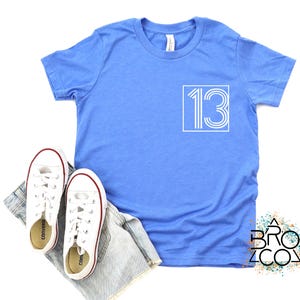 Op de afbeelding: Een lichtblauw t-shirt met het nummer 13 in een witte vierkant. Witte sneakers met rode rand en een spijkerbroekshort liggen naast het shirt. Het shirt heeft een ronde hals en een label.