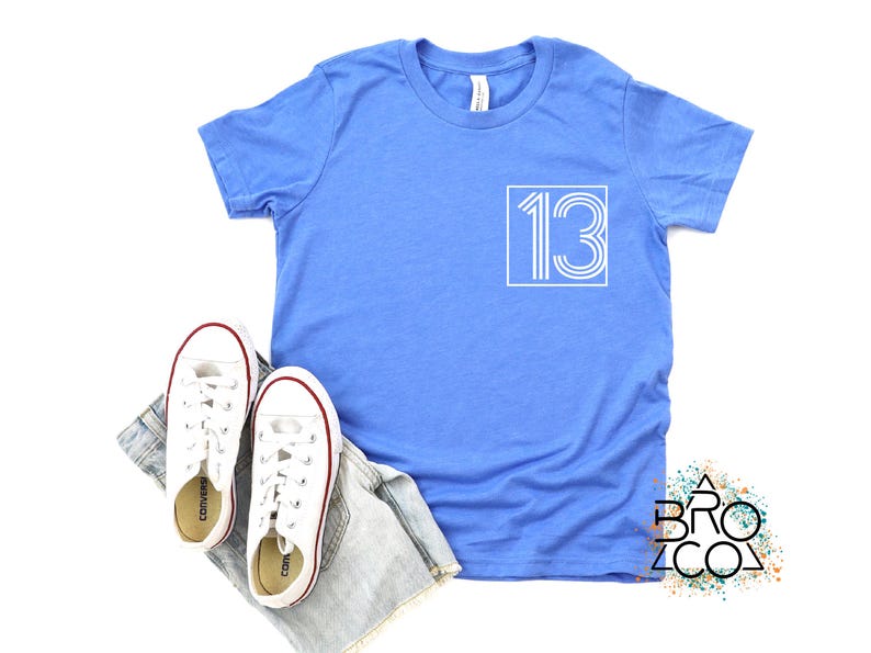 Op de afbeelding: Een lichtblauw t-shirt met het nummer 13 in een witte vierkant. Witte sneakers met rode rand en een spijkerbroekshort liggen naast het shirt. Het shirt heeft een ronde hals en een label.