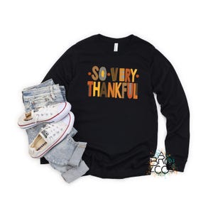 May include: A black long-sleeve shirt featuring the text SO VERY THANKFUL in orange, yellow, and brown block letters, paired with light blue denim jeans and white canvas sneakers with red and blue stripes on the rubber soles.