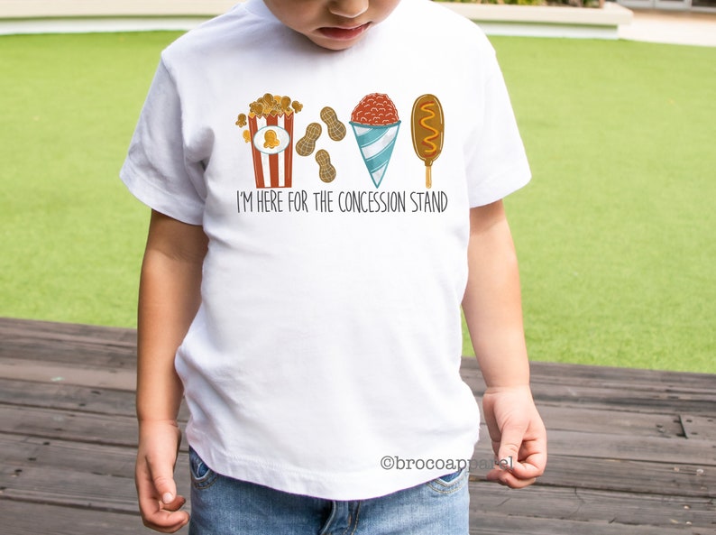 May include: White t-shirt featuring a graphic of popcorn, peanuts, a snow cone, and a corn dog. The text on the shirt reads "I'm here for the concession stand."