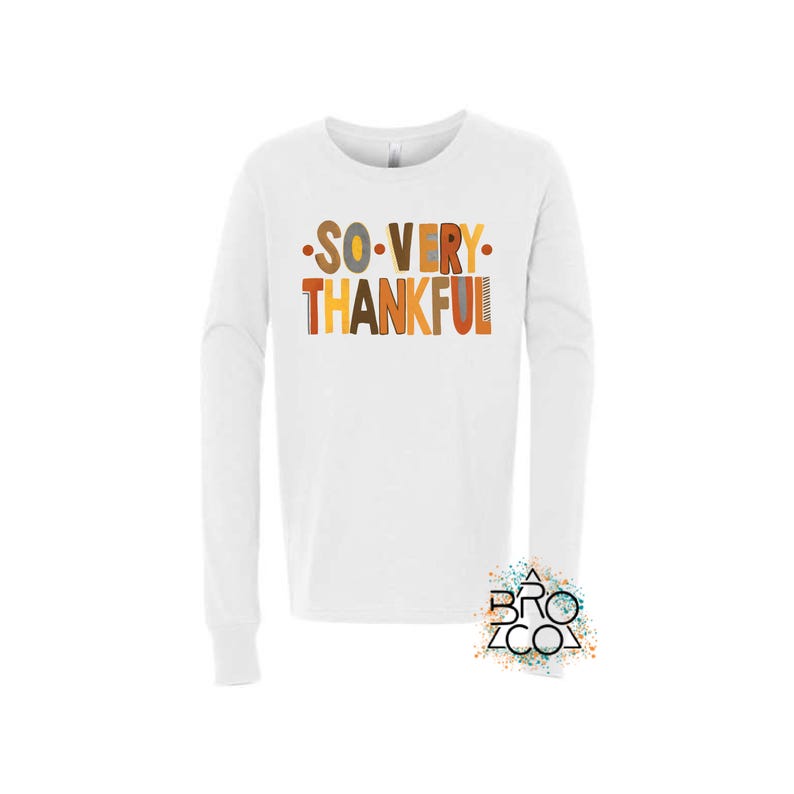 May include: A white long-sleeve shirt featuring the text SO VERY THANKFUL in a multi-colored palette of orange, brown, and yellow. The letters have varied textures and patterns, and the shirt includes a small logo in the bottom right corner.