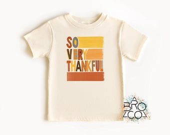May include: Cream-colored toddler t-shirt with the words "SO VERY THANKFUL" in colorful, block letters. The text is layered over horizontal brushstroke-style stripes in yellow, orange, and brown. The shirt has short sleeves and a crew neck.