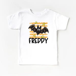 May include: A white short-sleeve t-shirt featuring a graphic of a black bat with white eyes, surrounded by yellow stars and horizontal orange stripes. The name FREDDY is printed in bold black capital letters underneath the bat illustration.