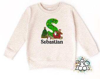 May include: A beige sweatshirt featuring a green letter "S" decorated with Christmas lights. The design includes a Christmas tree, Santa Claus in a sleigh, and the name "Sebastian" in black text. A festive holiday-themed garment.