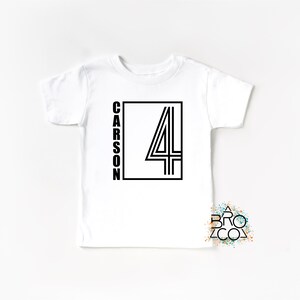May include: A white short-sleeved t-shirt featuring a black graphic design on the front. The design consists of a rectangular frame containing the number 4 and the name CARSON written vertically in a bold, sans-serif black font.