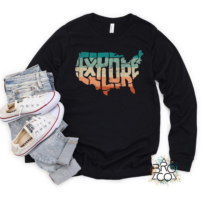May include: A black long-sleeve t-shirt featuring a graphic of the United States map filled with the word EXPLORE in a distressed, multi-colored retro font. The shirt is paired with light blue denim jeans and white canvas sneakers.