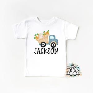 May include: White t-shirt with a blue truck carrying a basket of carrots and a bunny. The text "JACKSON" is printed below the truck.