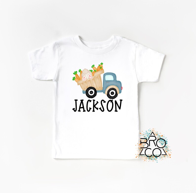 May include: White t-shirt with a blue truck carrying a basket of carrots and a bunny. The text "JACKSON" is printed below the truck.