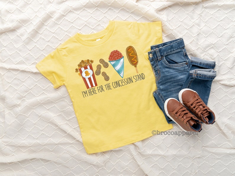 May include: Yellow t-shirt with a graphic of popcorn, peanuts, a snow cone, and a corn dog. The text on the shirt reads "I'm here for the concession stand."