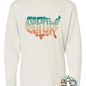 May include: A heathered cream long-sleeve shirt featuring a distressed graphic of the United States map filled with horizontal stripes in teal, tan, and orange. The word EXPLORE is integrated into the map design. A logo with the text BRO CO is in the corner.