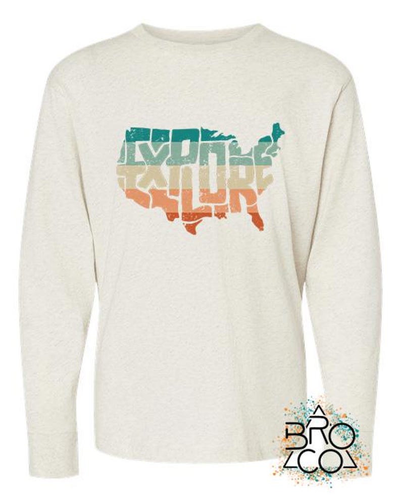 May include: A heathered cream long-sleeve shirt featuring a distressed graphic of the United States map filled with horizontal stripes in teal, tan, and orange. The word EXPLORE is integrated into the map design. A logo with the text BRO CO is in the corner.