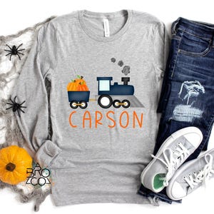 May include: A gray long-sleeve shirt with a train graphic carrying pumpkins and the name "Carson" in orange. The image includes a pair of blue jeans and gray sneakers, with Halloween decorations.