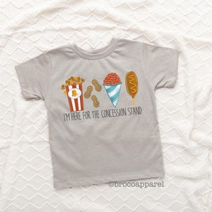 May include: A grey t-shirt with a graphic of popcorn, peanuts, a snow cone, and a corn dog. The text "I'm here for the concession stand" is printed below the graphic.