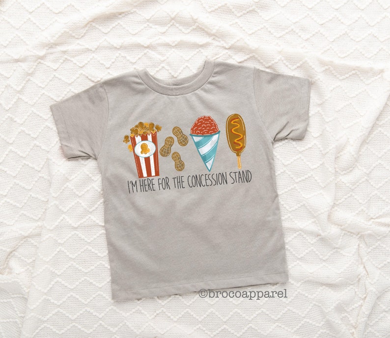 May include: A grey t-shirt with a graphic of popcorn, peanuts, a snow cone, and a corn dog. The text "I'm here for the concession stand" is printed below the graphic.