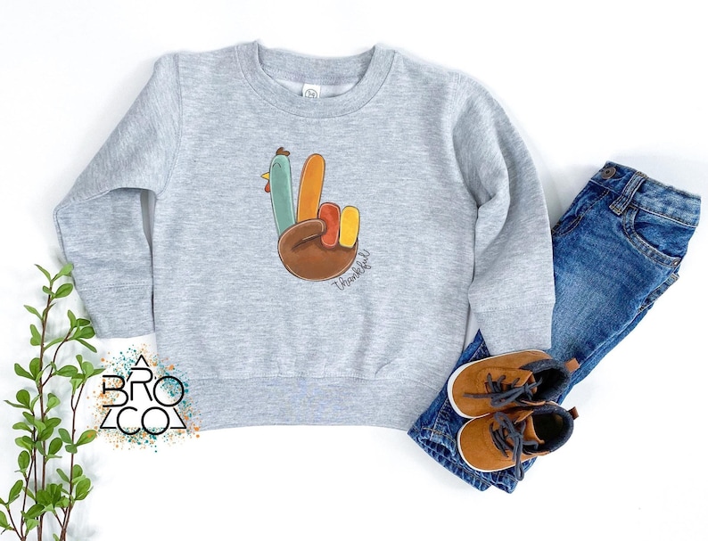 Może przedstawiać: Jasnoszara bluza z kolorowym gestem dłoni indyka i napisem "Thankful". Projekt zawiera brązowy, pomarańczowy, ż&oacute;łty, zielony i niebieski. Bluza jest zestawiona z niebieskimi dżinsami i brązowymi butami.