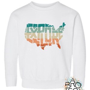 May include: A white long-sleeve sweatshirt featuring a distressed graphic of the United States map filled with the word EXPLORE in a retro-style font. The graphic uses horizontal stripes in teal, beige, and orange colors across the map shape.