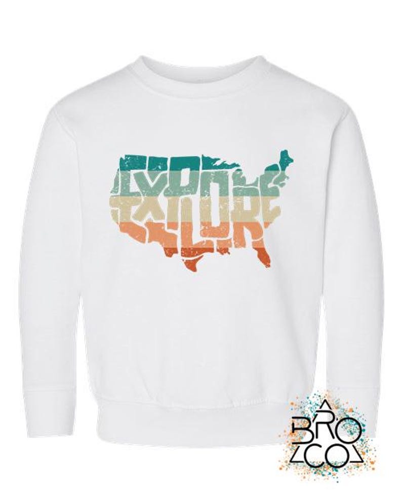 May include: A white long-sleeve sweatshirt featuring a distressed graphic of the United States map filled with the word EXPLORE in a retro-style font. The graphic uses horizontal stripes in teal, beige, and orange colors across the map shape.