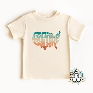 May include: A cream-colored short-sleeve t-shirt featuring a graphic of the United States map filled with the word EXPLORE in a distressed, multi-colored horizontal striped pattern of teal, beige, and orange. A logo with the text BRO CO is visible.