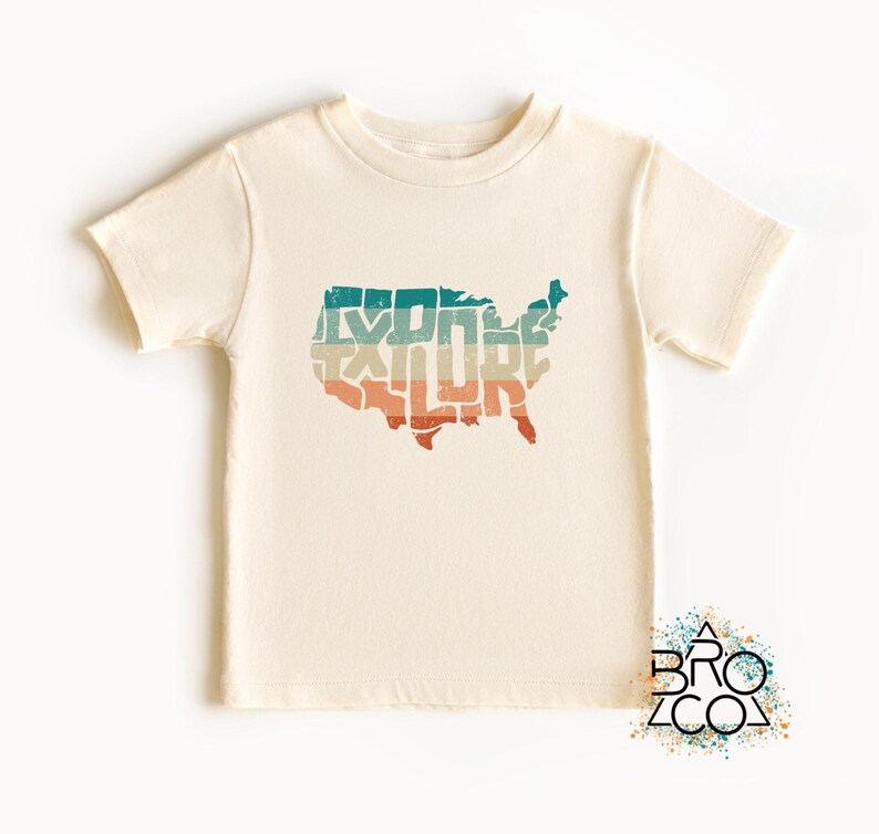 May include: A cream-colored short-sleeve t-shirt featuring a graphic of the United States map filled with the word EXPLORE in a distressed, multi-colored horizontal striped pattern of teal, beige, and orange. A logo with the text BRO CO is visible.