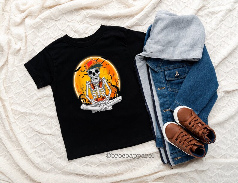 May include: Black t-shirt featuring a skeleton wearing a cap and headphones, set against an orange moon backdrop with bats and trees. The skeleton is in a seated position. A denim jacket, gray hoodie, and brown sneakers are next to the shirt. The text "brocoapparel" is at the bottom.