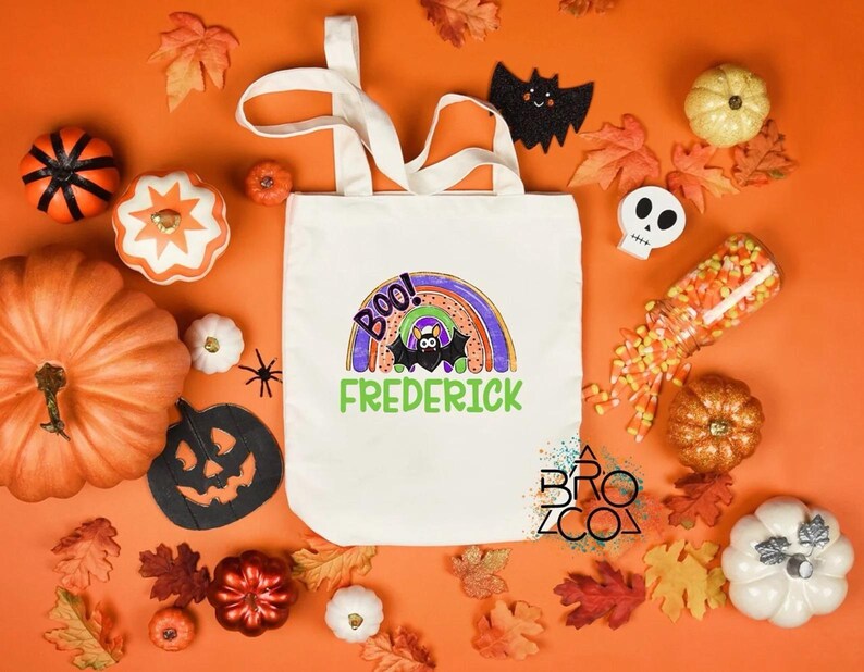 Puede incluir: Un bolso de mano blanco con un dise&ntilde;o tem&aacute;tico de Halloween, que incluye un arco&iacute;ris, un murci&eacute;lago y la palabra "BOO!" y el nombre "Frederick" en verde. La imagen incluye calabazas, hojas y caramelos de ma&iacute;z sobre un fondo naranja.