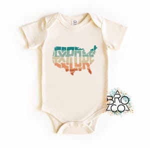 May include: A cream-colored short-sleeve baby bodysuit featuring a distressed graphic of the United States map filled with the word EXPLORE in horizontal stripes of teal, beige, and orange. A logo with the text BRO CO is visible.